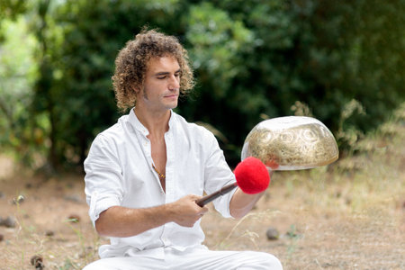 A man playing on a Tibetan singing bowls sitting outdoors for meditation. Buddhist healing practices. Sound therapy, alternative medicine. Clearing the space of negative energy. selective focusの写真素材
