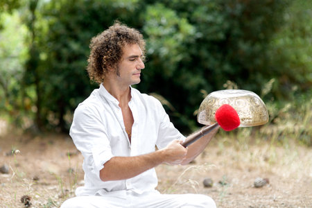 A man playing on a Tibetan singing bowls sitting outdoors for meditation. Buddhist healing practices. Sound therapy, alternative medicine. Clearing the space of negative energy. selective focusの写真素材