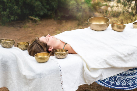 Young woman laying on a massage bed with tibetan singing bowl outdoor. Nepal Buddha singing bowls sound massage therapy is Buddhist healing practices. Caucasian woman relax with alternative medicineの写真素材