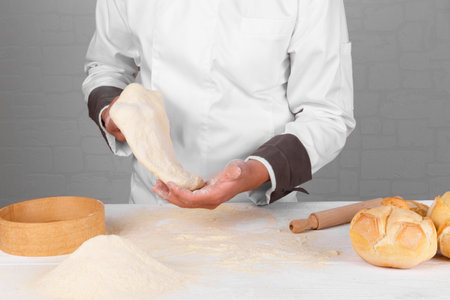 A baker holding bread dough in his hands, next to fresh Italian wheat bread on a table with flour on...