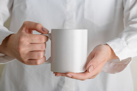 Female hand holding white mug mockup with blank copy space for your advertising text message or promotional content. Girl in white shirt holding white porcelain coffee mug mock upの写真素材