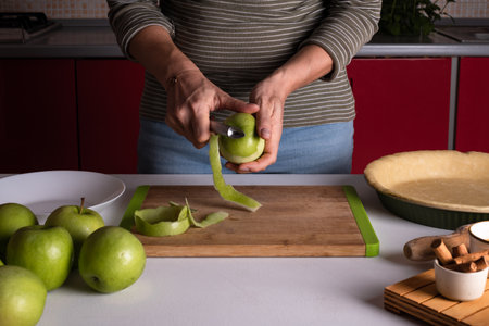 Woman is peeling apples to prepare apple pie in the kitchen. Authentic female hands peel an apple with a knife. Cooking apple pie process, recipe step by step. Thanksgiving preparation, autumn bakeryの写真素材