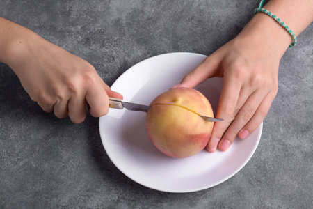 Authentic female hands cutting a fresh ripe peach on white plate on a gray table background, close up. Healthy summer snack, food lifestyleの写真素材