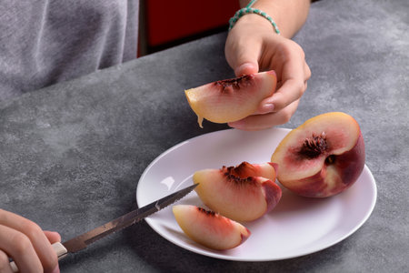 Authentic female hands holding a piece of a fresh ripe peach on white plate on a gray table...