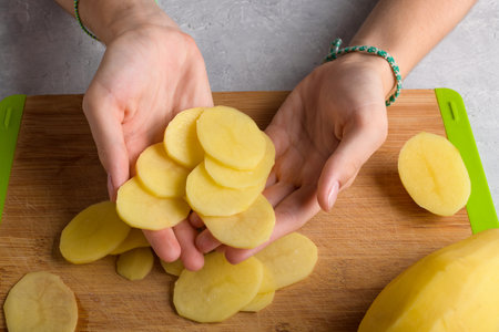 Authentic female hands is holding cutting potatoes on wooden cutting board on kitchen table. Woman in apron cut potato, preparing food, home interior, domestic life, lifestyle, top viewの写真素材