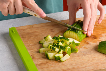 Authentic female hands cutting zucchini by cubes on wooden cutting board on kitchen table