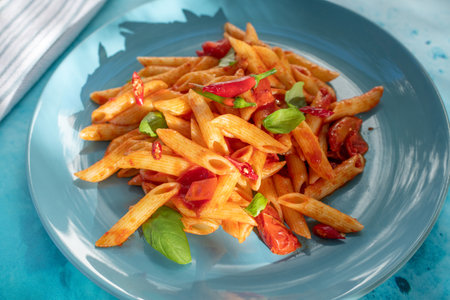 Spicy Italian penne all'Arrabbiata pasta on blue ceramic plate with tomato chili sauce on blue kitchen table,close upの写真素材