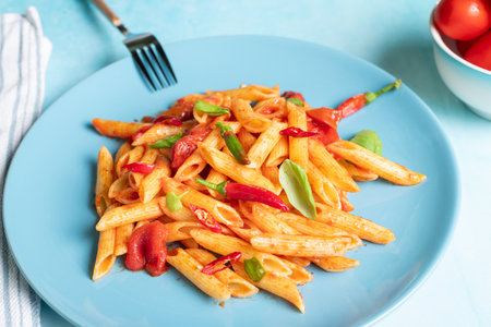 Classic penne all'Arrabbiata, spicy pasta with tomato and chilli sauce, on ceramic blue plate on blue kitchen table on sunny dayの写真素材