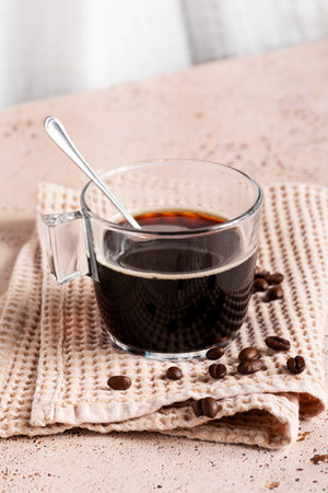 Glass cup of strong soluble black coffee on waffle cloth with roasted coffee beans on textured napkin on beige stone kitchen table, Coffee break, refreshing energy drinkの写真素材