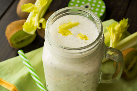 green smoothie in a glass jar with lid and a straw, celery and kiwi over dark wooden tableの写真素材