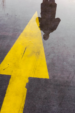Yellow arrow, yellow straight sign on wet pavement, direction arrow. Reflections of a man in the...