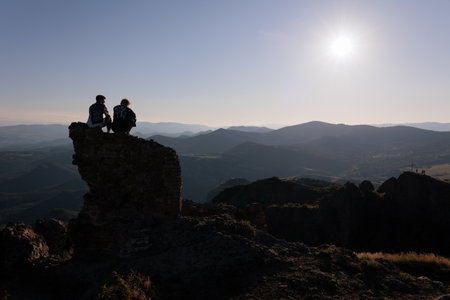 Silhouette of a couple at sunset overlooking the mountains. 2 friends are sitting on top of a permitted brick wall in the sky and mountains. Kojori, Georgia. horizontal photo, high contrast photoの写真素材