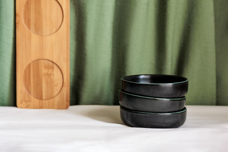 Minimalist composition with stacked black bowls, a wooden tray, and a green fabric backdrop