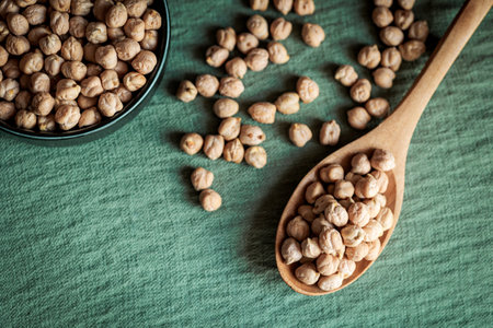 Top view of dried chickpeas in a ceramic bowl and a wooden spoon on a green background, showing rustic and healthy food conceptsの写真素材