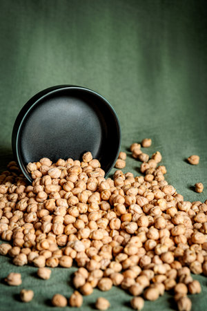 Black ceramic bowl tipped over, spilling raw chickpeas onto a green fabric surface. Natural light emphasizes the warm tones and textured details of the legumes, creating a rustic, organic aestheticの写真素材