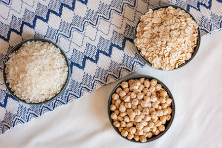 Three ceramic bowls filled with rice, oats, and chickpeas placed on a white surface with a textured...