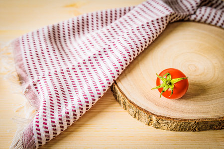 A single cherry tomato on a wooden cutting board with a checkered towel. minimalist food composition highlighting organic produce, rustic aesthetics, and natural ingredients in a cozy kitchen settingの写真素材