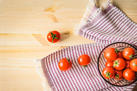 A bowl of cherry tomatoes on a wooden table with a checkered towel, with some scattered around. A rustic food composition highlighting freshness, organic produce, and natural ingredients for cookingの写真素材