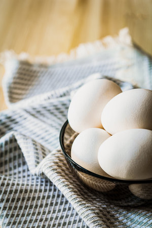 A close-up of fresh white eggs in a black glass bowl on a rustic wooden table with a textured linen napkin. Soft natural light creates a cozy atmosphere, perfect for organic food themesの写真素材