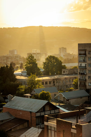 Golden sunlight streaming through clouds over Tbilisi cityscape, casting dramatic light beams over residential buildings and trees at sunset. Warm, atmospheric urban viewの写真素材