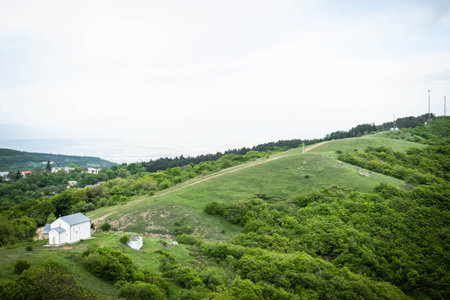 Small white church on a green hillside overlooking a distant city, with a forest in the background. Peaceful rural scene in Georgiaの写真素材