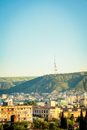 Vertical panorama of Tbilisi, Georgia, with Mtatsminda hill and iconic TV tower towering over the city under a clear blue sky. A mix of modern and Soviet-era buildingsの写真素材