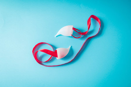 White menstrual cups with red ribbon on a blue background, shaped like awareness symbols. Creative visual for eco-friendly period care and menstrual health awarenessの写真素材