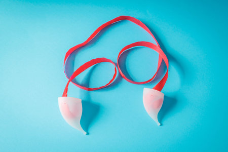 White menstrual cups with red ribbon on a blue background, shaped like awareness symbols. Creative visual for eco-friendly period care and menstrual health awarenessの写真素材