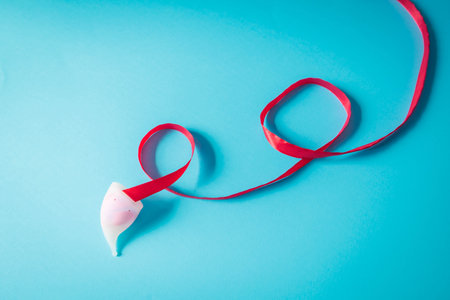 Minimalist composition of a white menstrual cup with flowing red ribbon on blue backdrop. Symbolic image promoting feminine hygiene and eco-conscious menstruationの写真素材