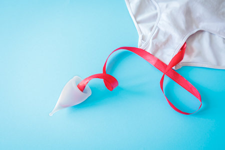 Menstrual cup with red ribbon symbolizing period blood, placed next to white underwear on pastel blue background. Feminine hygiene concept with clean and minimal aestheticの写真素材