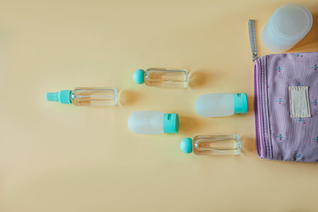 Flat lay of travel-size toiletry bottles and a purple cosmetic pouch on a pastel yellow background....