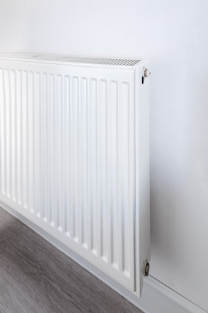 Angled view of a white radiator with visible valve and slotted grill, showing heating appliance...