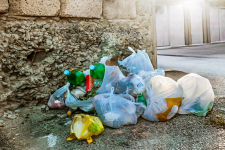 Pile of plastic garbage bags and bottles dumped near a rough stone wall on an urban street corner, highlighting issues of waste management and environmental pollution.の写真素材