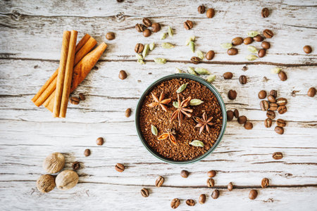 Ground coffee in rustic bowl garnished with star anise and cardamom, framed by cinnamon sticks, nutmeg and scattered beans on weathered woodの写真素材