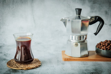 Curved glass of freshly brewed coffee on woven coaster opposite a moka pot and bowl of roasted beans on bamboo board, wide layout with roomy blank space for headline copyの写真素材