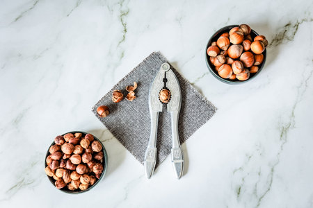 Symmetrical flat-lay: twin bowls of whole and shelled hazelnuts flank ornate metal nutcracker on...