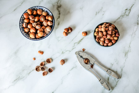 Rustic flat-lay of whole and shelled hazelnuts with vintage metal nutcracker on white marble countertop, shot in natural daylight, airy Scandinavian kitchen moodの写真素材
