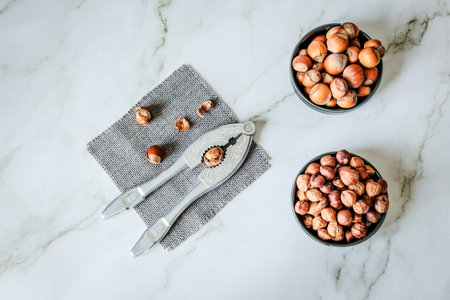 Overhead layout of hazelnuts in two ceramic bowls beside vintage nutcracker on gray woven cloth over...