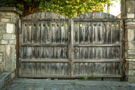 Large old wooden double gates with iron rings and circular cut-outs, framed by stone pillars in Mtskheta, Georgia. Aged texture and traditional craftsmanship in natural lightの写真素材