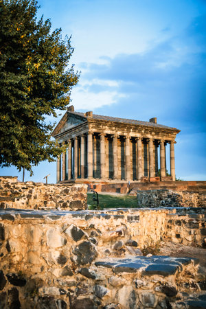 Vertical view of the Temple of Garni in Armenia with foreground ruins and a large tree. Captured at sunset, highlighting the templeの写真素材