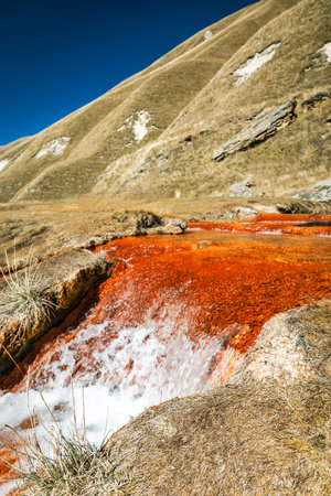 Vivid red mineral spring in Truso Valley, Georgia, during autumn. A surreal natural stream rich in iron colors flows through dry mountain slopes under a clear blue skyの写真素材