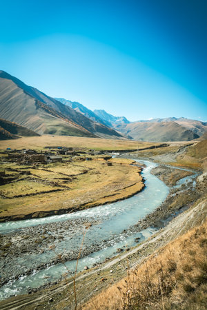 Aerial view of the abandoned Ossetian village in Truso Valley, Georgia. A winding river flows through golden fields and majestic mountains under a clear autumn skyの写真素材