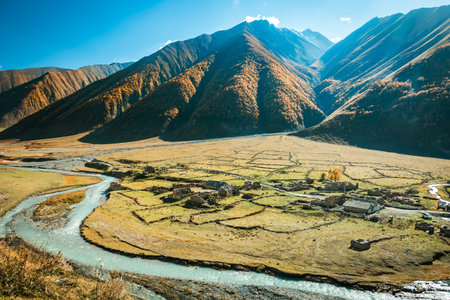 Aerial view of the abandoned Ossetian village in Truso Valley, Georgia. A winding river flows through golden fields and majestic mountains under a clear autumn skyの写真素材