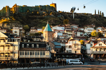 Panoramic morning view of Tbilisi with the Narikala Fortress, cable cars in the sky, and traditional houses illuminated by soft golden sunrise light over the old districtの写真素材