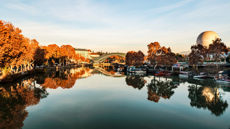Panoramic autumn scene of the Kura River in Tbilisi with colorful trees, the iconic Peace Bridge, and a cable car crossing the sky, all bathed in warm evening lightの写真素材