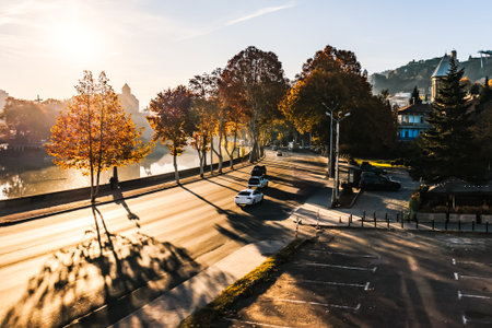 Morning sunlight casts long shadows over a quiet riverside street in Tbilisi, Georgia. Autumn trees...