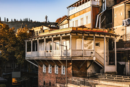Traditional wooden house with carved balcony in Tbilisi, Georgia, illuminated by golden morning light. Autumn leaves frame the warm scene, showcasing the city's old charmの写真素材