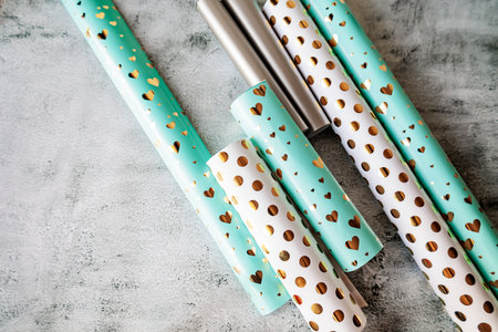 Top view of colorful wrapping paper rolls in gold heart and polka dot patterns on neutral background. Modern festive flat lay for gift packaging themesの写真素材