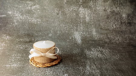 White candle wrapped in rustic burlap and decorated with beige ribbon, placed on woven mat over dark textured background. Minimalist cozy decor for holidays, gift or relaxationの写真素材