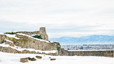 Ancient snow-covered walls and stone ruins above Kutaisi, Georgia, with dramatic mountain landscape and modern city buildings below. Winter view blending history and urban natureの写真素材